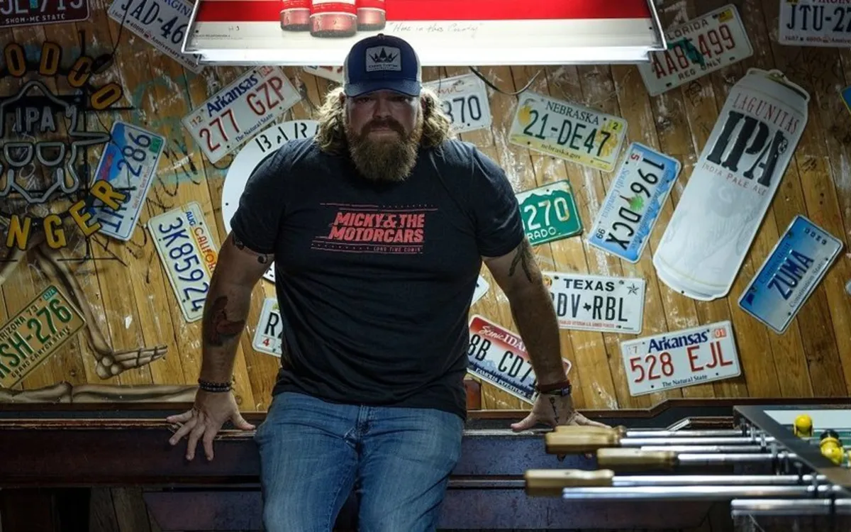 Drew Cooper in a ball cap and beard, sitting in front of a wall of license plates