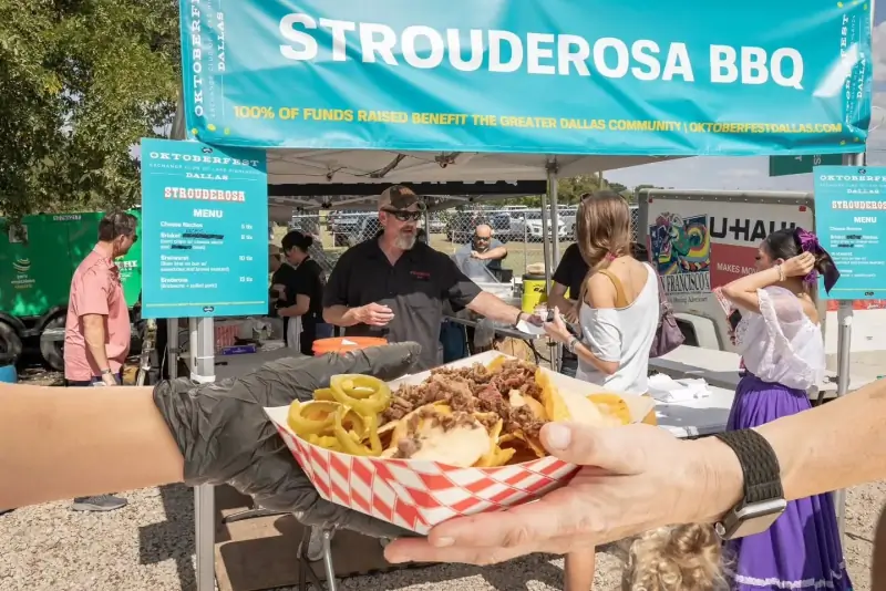A hand holds up a tray of brisket nachos with pickled jalapeños in front of the Strouderosa BBQ tent at Oktoberfest Dallas, the menu board and a line of customers behind it